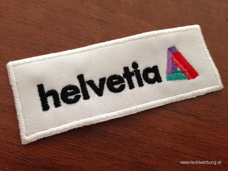 Rectangular white embroidered patch with a black company name and a small multi-coloured triangle symbol, lying on a wooden table.